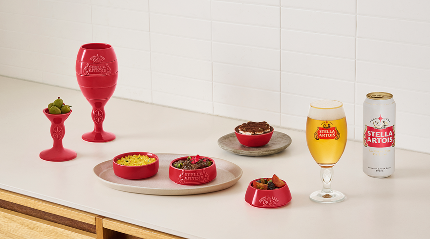 스텔라 아르투아의 빨간 고메 챌리스에 음식이 담겨있으며, 스텔라 챌리스 전용잔과 500ml 캔 맥주가 테이블에 놓여있습니다.